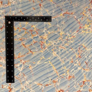 May include: A black right-angle ruler rests on a marbled paper surface. The paper has a light blue background with white, gold, and red veining. The ruler is marked in inches, with measurements up to 7 inches.