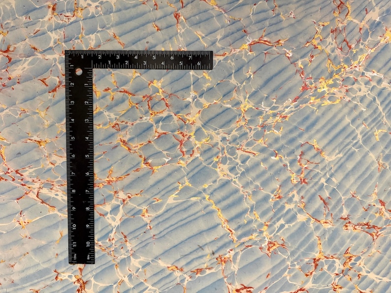 May include: A black right-angle ruler rests on a marbled paper surface. The paper has a light blue background with white, gold, and red veining. The ruler is marked in inches, with measurements up to 7 inches.