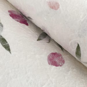 May include: Close-up of handmade paper with a floral design. The paper is white with pink flower petals and green leaves. The paper is rolled up, showing the texture and layers of the material.