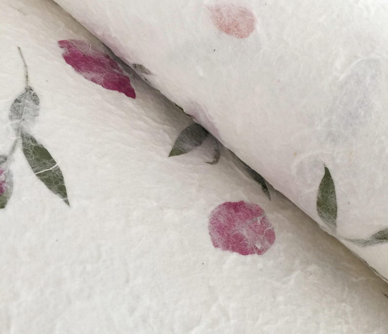 May include: Close-up of handmade paper with a floral design. The paper is white with pink flower petals and green leaves. The paper is rolled up, showing the texture and layers of the material.