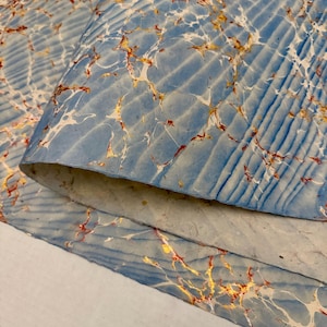 May include: Close-up of marbled paper with a blue background and white, gold, and red veining. The paper has a textured, ridged pattern. The edges of the paper are visible, showing the layers and texture.