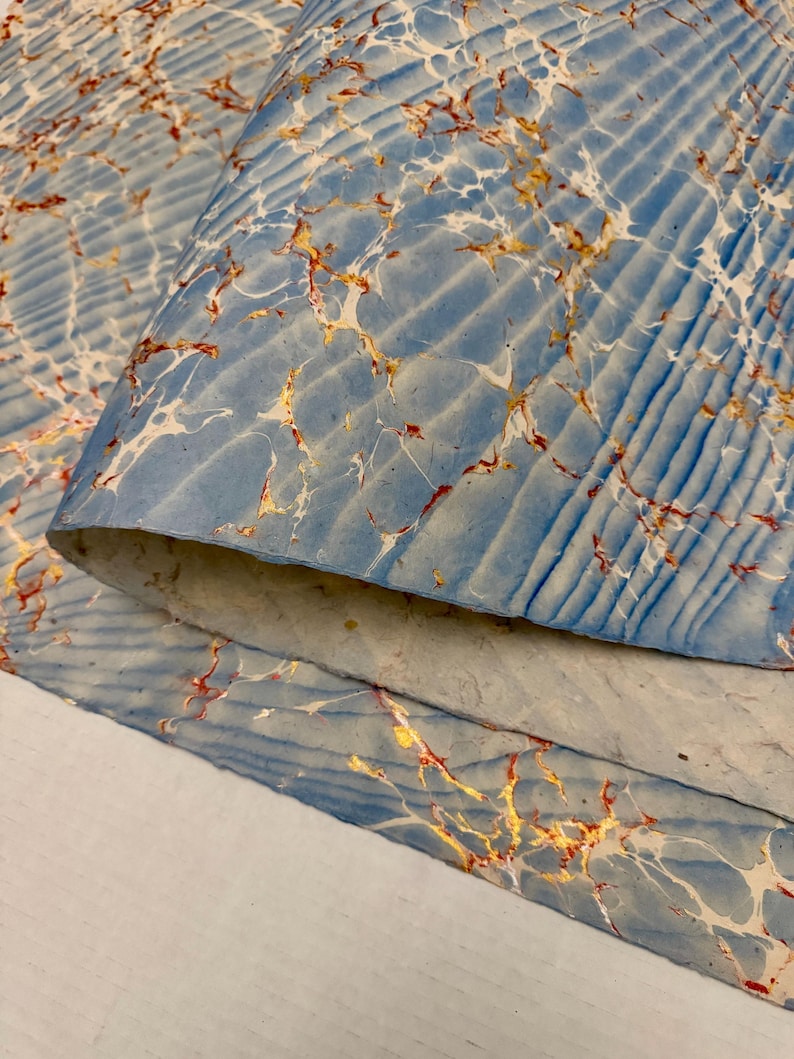 May include: Close-up of marbled paper with a blue background and white, gold, and red veining. The paper has a textured, ridged pattern. The edges of the paper are visible, showing the layers and texture.