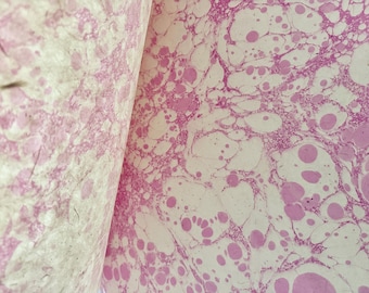 May include: Close-up of marbled paper with a pink and white pattern. The design features swirling, organic shapes and circular patterns. The paper is folded, revealing the pattern on both sides.