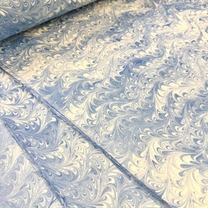 May include: Close-up of a stack of marbled paper with a swirling pattern in shades of blue and cream. The paper has a textured appearance, with the design resembling flowing water or feathers. The paper is likely used for stationery or art projects.