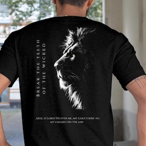 Op de afbeelding: Zwart T-shirt met een grijswaarden illustratie van een leeuwenprofiel. De tekst "BREAK THE TEETH OF THE WICKED" staat verticaal links. Daaronder staat: "ARISE, O LORD! DELIVER ME, MY GOD! STRIKE ALL MY ENEMIES ON THE JAW:"