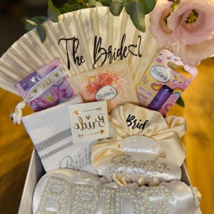 Könnte beinhalten: Eine Geschenkbox mit Brautaccessoires, darunter ein Fächer mit der Aufschrift "The Bride", ein Satin-Bademantel, Socken und andere Artikel. Die Box ist mit Blumen und Grün dekoriert.