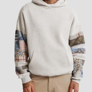 May include: A light gray hoodie with patchwork sleeves. The sleeves feature a mix of patterned fabrics in shades of blue, brown, and floral prints. The hoodie has a front pocket and a drawstring hood.