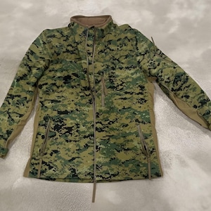 May include: A full-zip jacket with a digital camouflage pattern in shades of green, black, and tan. The jacket features a stand-up collar, two zippered pockets, and a tan-colored lining along the sides and sleeves.