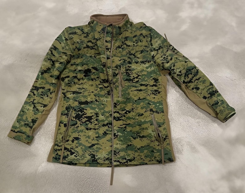 May include: A full-zip jacket with a digital camouflage pattern in shades of green, black, and tan. The jacket features a stand-up collar, two zippered pockets, and a tan-colored lining along the sides and sleeves.
