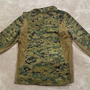 May include: A long-sleeved camouflage shirt in a digital pattern of green, brown, and tan. The shirt has a zipper at the neck and tan-colored panels on the sides and sleeves. The garment is designed for outdoor activities.