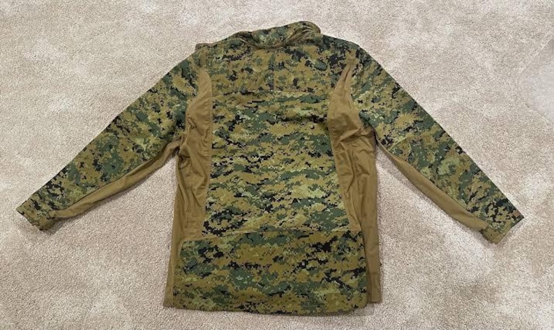 May include: A long-sleeved camouflage shirt in a digital pattern of green, brown, and tan. The shirt has a zipper at the neck and tan-colored panels on the sides and sleeves. The garment is designed for outdoor activities.
