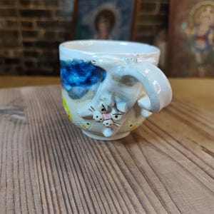 May include: A ceramic mug with a quirky cat design. The mug features a white cat with a grumpy expression, a curved handle, and a painted landscape with blue and yellow accents. The mug is on a wooden surface.