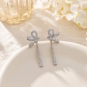 May include: A pair of silver-toned drop earrings featuring a bow-shaped stud encrusted with small clear stones. A vertical line of round clear stones hangs from each bow, creating a dangling design that catches the light with a metallic finish.