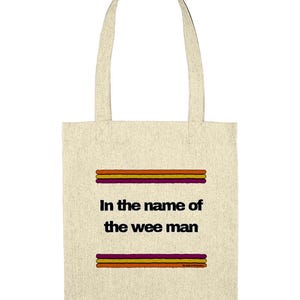 May include: Cream-colored tote bag with the text "In the name of the wee man" in black. The text is between two sets of horizontal stripes in orange, yellow, and purple. The bag has matching cream-colored handles.