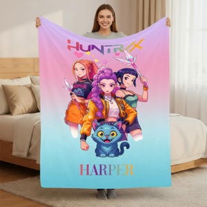 Puede incluir: Una manta colorida con una ilustración de dibujos animados de tres personajes de anime y un gato azul. La manta tiene un fondo degradado de rosa, morado y azul. Las palabras "HUNTR" y "HARPER" están en la manta.