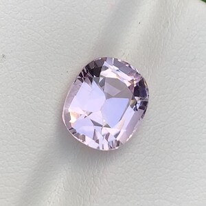 May include: A light purple, cushion-cut gemstone with a faceted surface. The gem is set on a white surface, with a blurred green background. The gemstone's facets reflect light, creating a sparkling effect. This gemstone is likely used in jewelry.