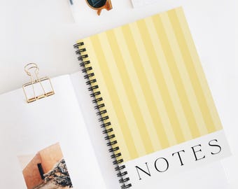 Op de afbeelding: Een spiraalgebonden notitieboek met een geel en lichtgeel gestreepte kaft. Het woord "NOTES" is in het zwart gedrukt op een witte band aan de onderkant. Een gouden paperclip en zonnebril zijn ook zichtbaar.