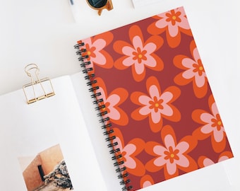May include: A spiral-bound notebook with a burgundy cover featuring a repeating pattern of orange and pink retro flowers. The notebook is open, revealing blank white pages and a gold paper clip.