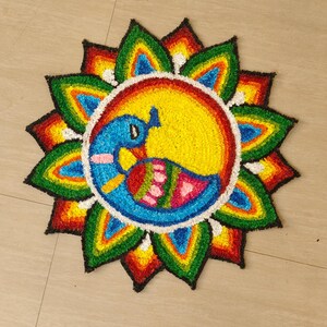 May include: A vibrant, circular floor decoration with a peacock motif. The peacock is rendered in blue, yellow, and pink, encircled by a white ring and a border of green, red, and yellow petals. The artwork is displayed on a light-coloured tiled floor.