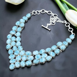 May include: A statement necklace featuring multiple oval-shaped, light blue and white marbled Larimar stones set in polished silver. The piece includes a silver chain with circular links and a toggle clasp closure, creating a wide, layered collar design.