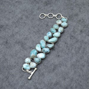 Könnte beinhalten: Ein silbernes Armband mit zwei Reihen unregelmäßiger, hellblauer Larimarsteine. Die Steine sind in Silber gefasst und durch eine Silberkette und einen Knebelverschluss verbunden. Das Armband wird auf einer grauen Oberfläche präsentiert.
