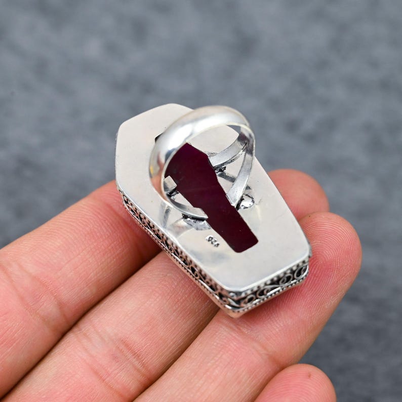 Large natural ruby faceted coffin cut deep vivid crimson red silver plated statement ring with double-layer oxidized filigree scrollwork and bead dot milgrain border setting, sleek polished silver band, held in hand against grey background