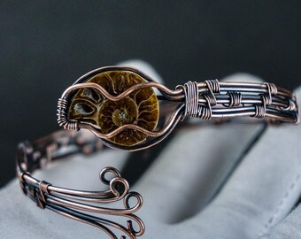 May include: A copper wire bracelet with a fossilised ammonite centrepiece. The ammonite is a spiral-shaped, brown and tan fossil. The bracelet has an open design with decorative swirls and woven wire detailing.