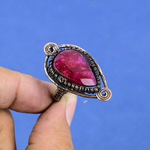 Puede incluir: Un anillo de color cobre con una gran piedra preciosa roja rubí en forma de lágrima. El anillo tiene un diseño calado adornado alrededor de la piedra y un detalle en espiral a cada lado. Fondo azul.