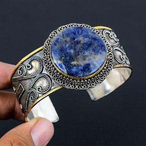 Könnte beinhalten: Ein silbernes Armreif-Armband mit einem großen, runden, blauen Sodalith-Stein in der Mitte. Der Stein ist von einer goldfarbenen Lünette und aufwendigem Silberfiligran umgeben. Das Armband hat ein breites Band mit dekorativen Schnörkeln.