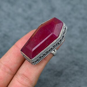 May include: A large, rectangular ruby gemstone ring. The ruby is a deep red colour and is set in a silver-coloured band with an ornate, filigree design around the stone. The ring is shown on a hand.