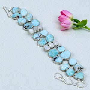 May include: A silver bracelet featuring multiple polished, light blue, oval-shaped Larimar stones interspersed with smaller, translucent white moonstones. The stones are set in silver bezels and connected in a multi-row design with a toggle clasp.
