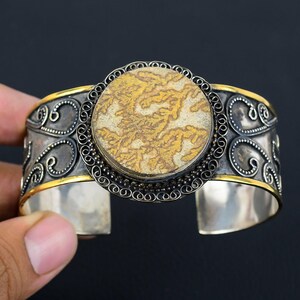 Könnte beinhalten: Silbernes Armreif-Armband mit einem großen, runden, gemusterten Stein in der Mitte. Der Stein ist hauptsächlich beige und goldfarben, eingefasst in einen dekorativen Silber- und Goldrahmen. Das Armband hat verschnörkelte Verzierungen und eine polierte Oberfläche.