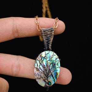 Può includere: Una collana con pendente artigianale con una conchiglia di abalone ovale e un design ad albero in filo metallico. Il pendente è sospeso a una catena color oro. La conchiglia di abalone mostra iridescenze blu, verdi e viola.