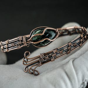 以下が含まれることがあります： 織り模様と緑色の石が特徴の銅線ブレスレット。ブレスレットは、複雑なワイヤーラッピングとユニークなオープンデザインが特徴です。石は楕円形で、濃い緑色をしています。ブレスレットは白い表面に表示されています。