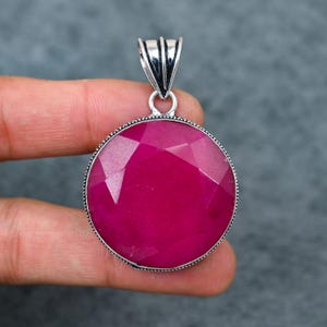 Può includere: Ciondolo rotondo con pietra preziosa magenta sfaccettata con un'anello argentato. La pietra è incastonata in una lunetta argentata con un bordo perlato. Il ciondolo è tenuto da una mano su sfondo grigio.