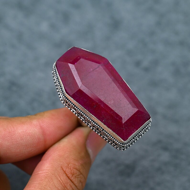 Large natural ruby faceted coffin cut deep vivid crimson red silver plated statement ring with double-layer oxidized filigree scrollwork and bead dot milgrain border setting, sleek polished silver band, held in hand against grey background