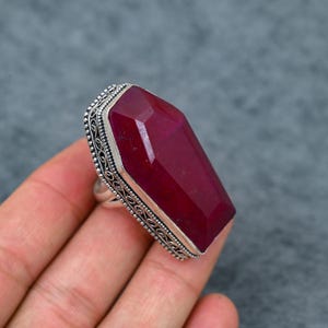 Large natural ruby faceted coffin cut deep vivid crimson red silver plated statement ring with double-layer oxidized filigree scrollwork and bead dot milgrain border setting, sleek polished silver band, held in hand against grey background