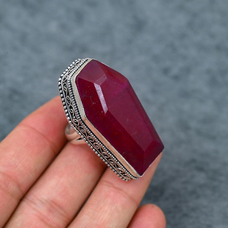 Large natural ruby faceted coffin cut deep vivid crimson red silver plated statement ring with double-layer oxidized filigree scrollwork and bead dot milgrain border setting, sleek polished silver band, held in hand against grey background