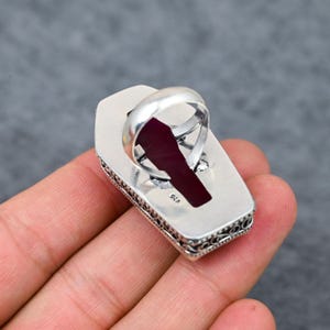 Large natural ruby faceted coffin cut deep vivid crimson red silver plated statement ring with double-layer oxidized filigree scrollwork and bead dot milgrain border setting, sleek polished silver band, held in hand against grey background