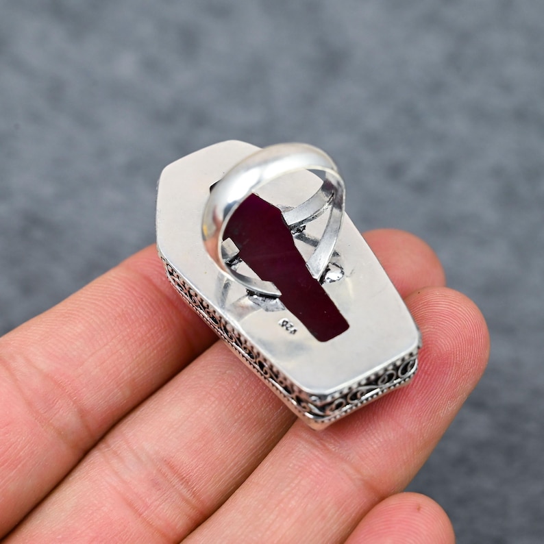 Large natural ruby faceted coffin cut deep vivid crimson red silver plated statement ring with double-layer oxidized filigree scrollwork and bead dot milgrain border setting, sleek polished silver band, held in hand against grey background