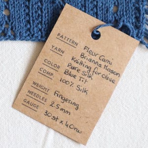 May include: A blue knitted swatch with fringe, featuring a delicate openwork design. A brown tag provides details: "Fleur Cami" pattern, "Blue Tit" color, and 100% silk composition. The gauge is 30 stitches x 40 rows.