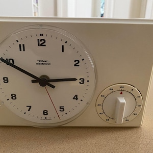 Può includere: Orologio elettronico Diehl color crema con quadrante rotondo e quadrante timer. Il quadrante dell'orologio ha numeri e lancette nere, con una lancetta dei secondi rossa. Il quadrante del timer è sul lato destro.