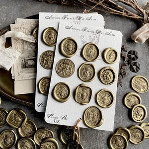 Puede incluir: Sellos de cera franceses antiguos dorados, con diseños en relieve detallados. Los sellos se presentan en tarjetas blancas con el texto "Antique French Wax Seals" y dispersos sobre una superficie gris.
