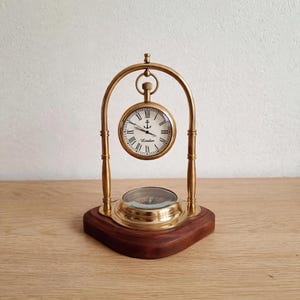 May include: A brass desk clock and compass set mounted on a dark wood base. The clock features a white face with black Roman numerals, an anchor icon, and the text London. The clock hangs from a brass arch above a circular brass compass.