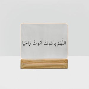 Könnte beinhalten: Rechteckiges, beleuchtetes Schild mit arabischer Schrift auf einem Holzsockel. Das Schild hat eine weiße Oberfläche mit schwarzem Text und einen warmen, weichen Schein an den Rändern. Der Holzsockel hat eine natürliche Holzmaserung.