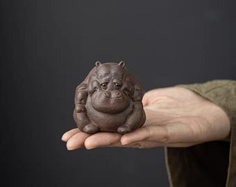 May include: A small, brown ceramic hippo figurine with a sad expression. The hippo is sitting upright with its arms crossed. The figurine is held in a person's hand against a dark background.