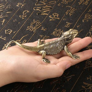 Könnte beinhalten: Eine detaillierte, bronzefarbene Eidechsenfigur mit strukturierter Haut und einem gekräuselten Schwanz. Das Reptil ist auf einer Hand positioniert und zeigt sein aufwendiges Design. Die Figur ist ein dekoratives Objekt.