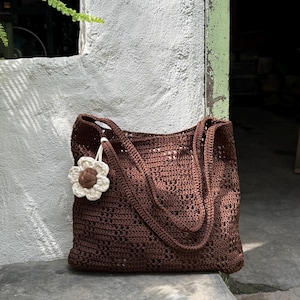 Puede incluir: Un bolso tote de ganchillo marrón con dos asas. El bolso tiene un charm de flor con centro marrón y pétalos blancos. El bolso está hecho de un material tejido y está sobre una superficie de hormigón.