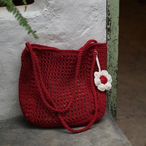 May include: A red crochet tote bag with long handles. The bag has a textured, open weave design. A white and red flower charm hangs from the side. The bag is sitting on a concrete surface.