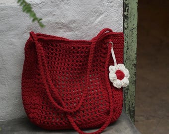 May include: A red crochet tote bag with long handles. The bag has a textured, open weave design. A white and red flower charm hangs from the side. The bag is sitting on a concrete surface.